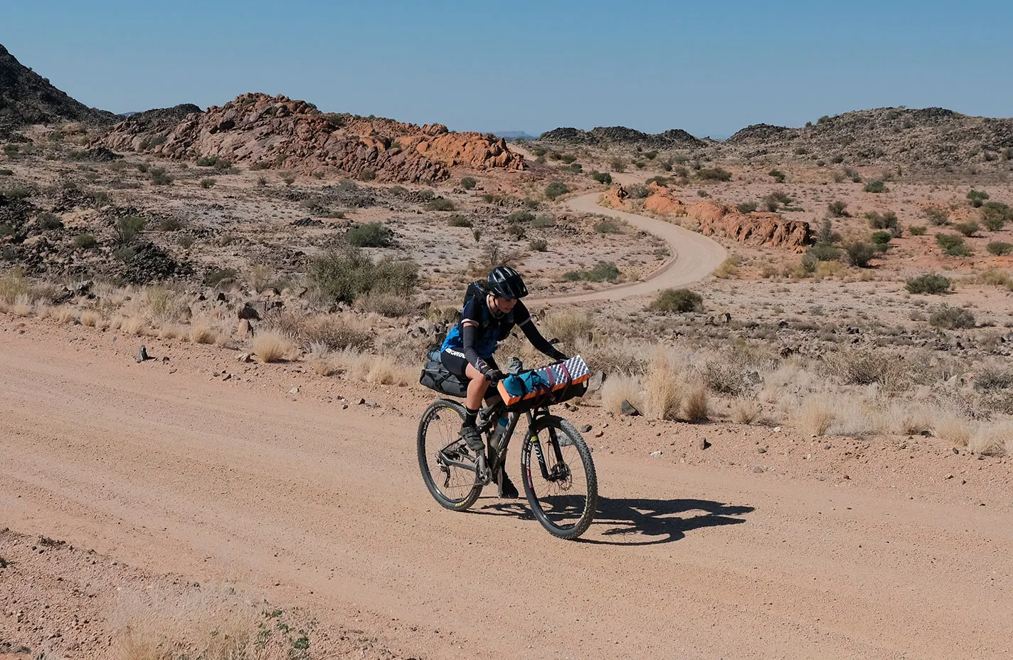 A cyclist riding a winding gravel road through arid, rocky hill country on a Wild Skies point-to-point route
